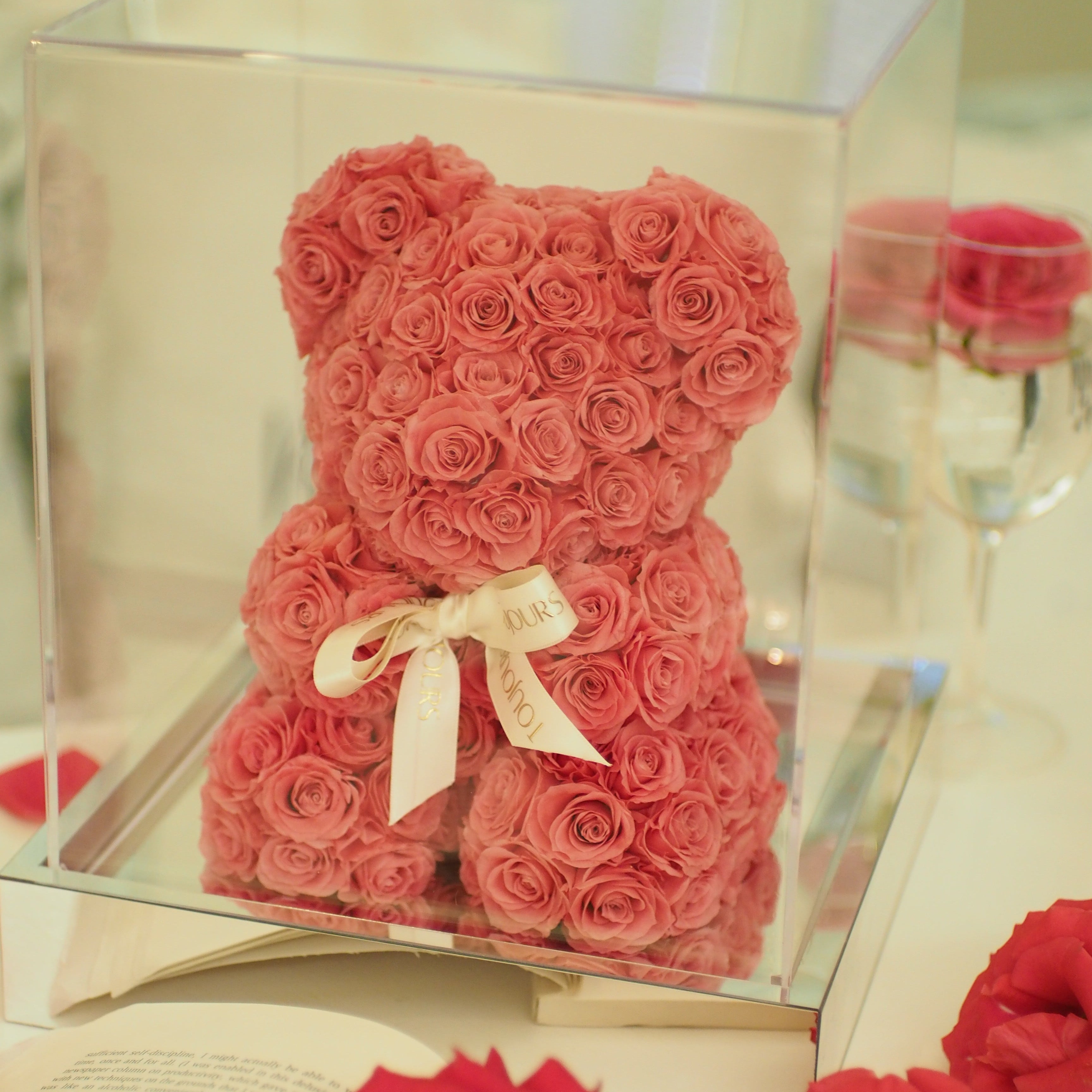 Teddy bear made of red roses in a clear box with a white ribbon on a reflective surface.