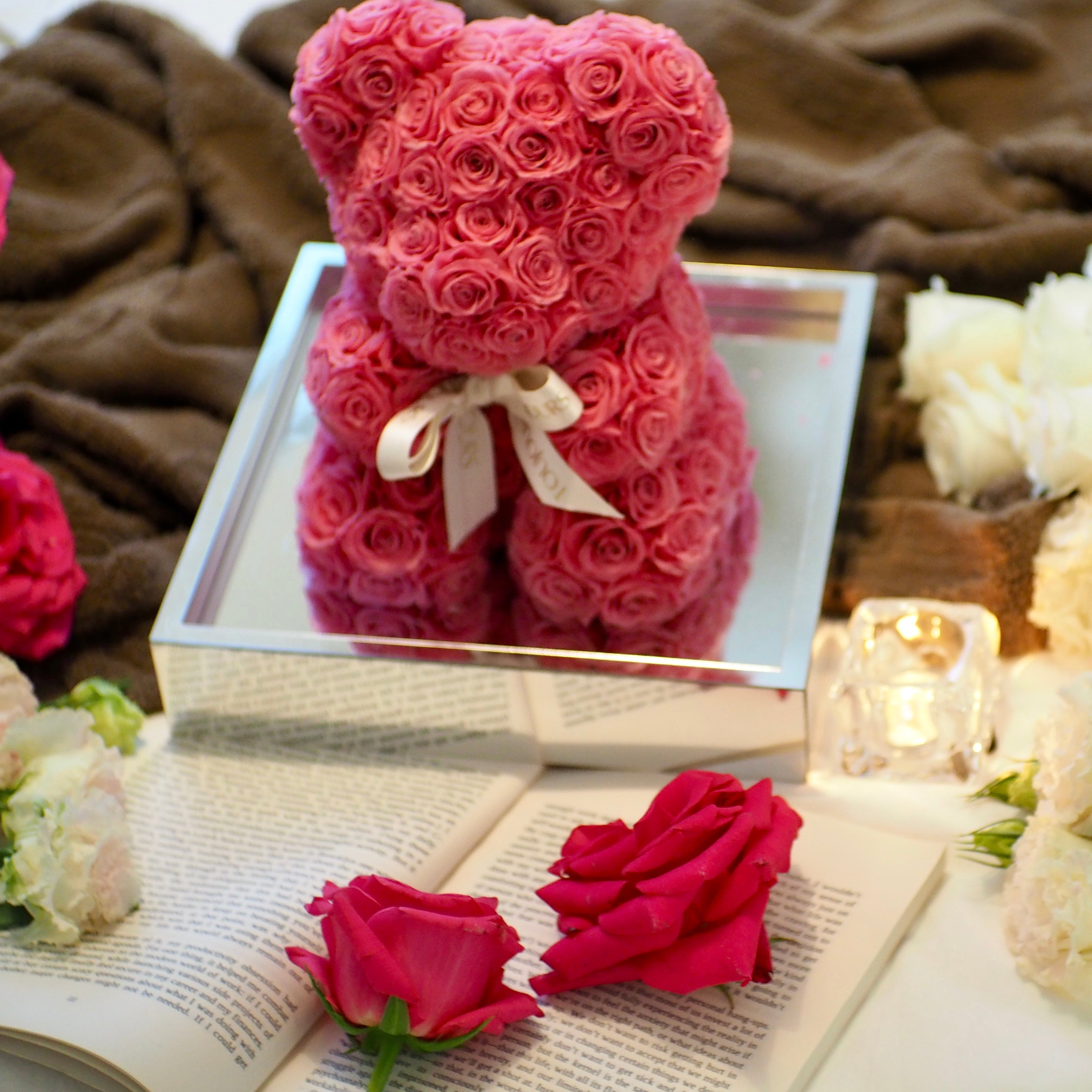 Pink teddy bear made of roses on a book with white and red roses around, on a brown fabric background.