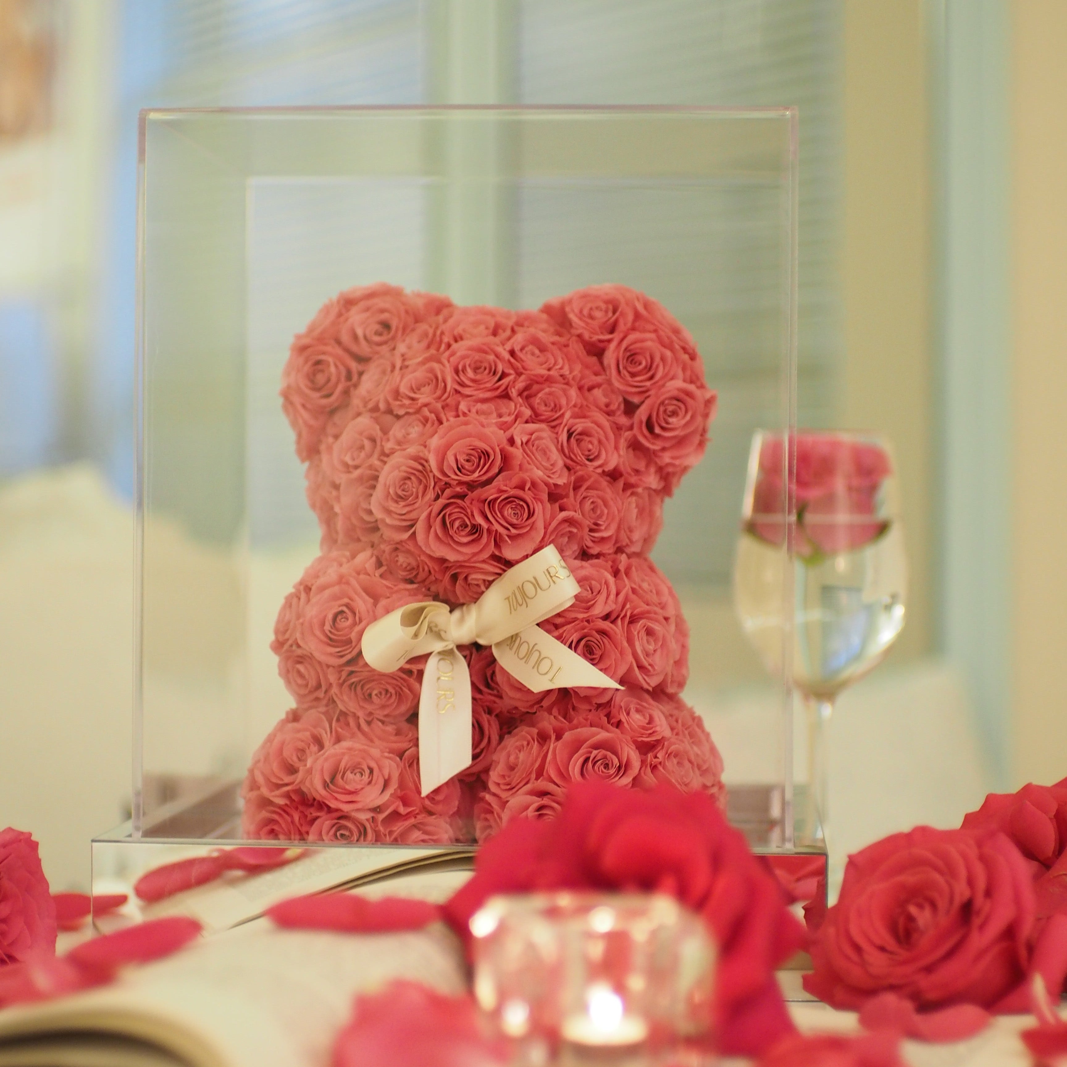 Red rose teddy bear with a bow in a clear box on a reflective surface