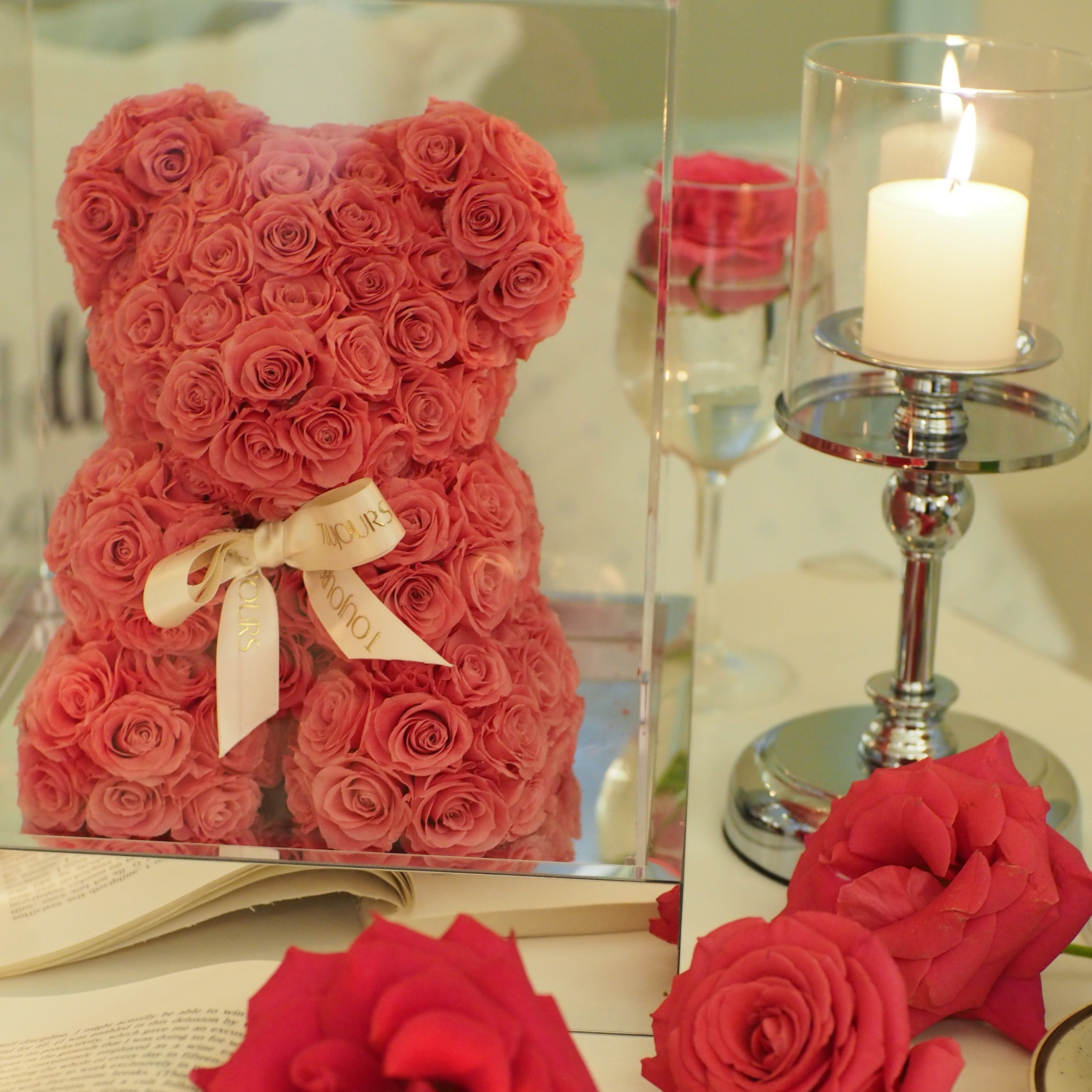 Red rose arrangement shaped like a bow with a decorative ribbon on a reflective surface.