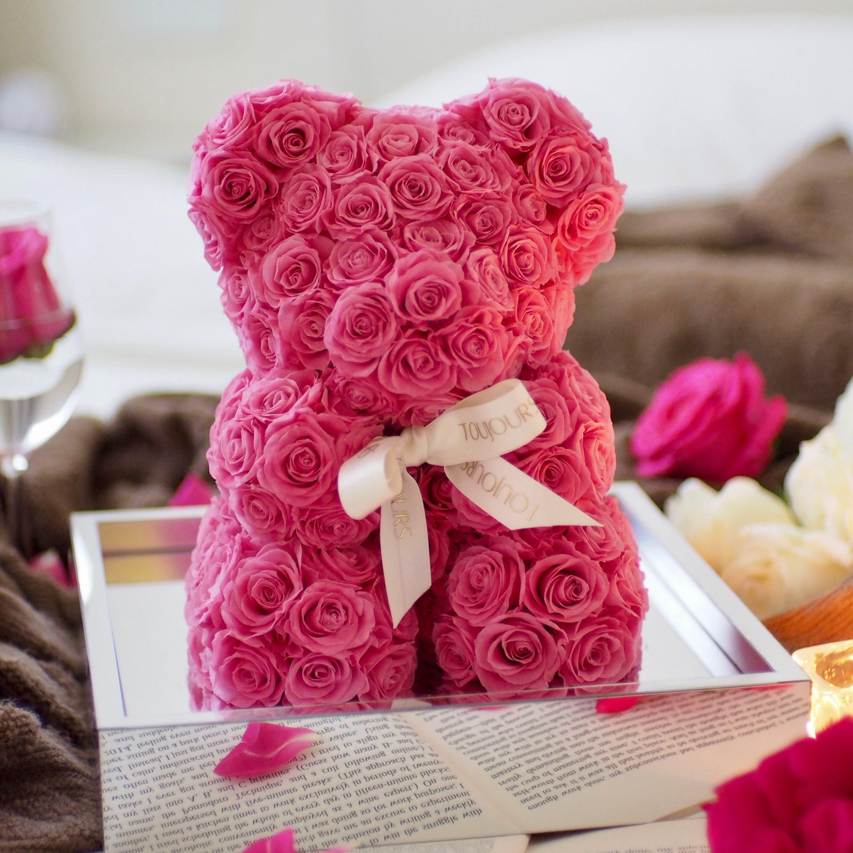Pink teddy bear made of roses with a white ribbon on a table.