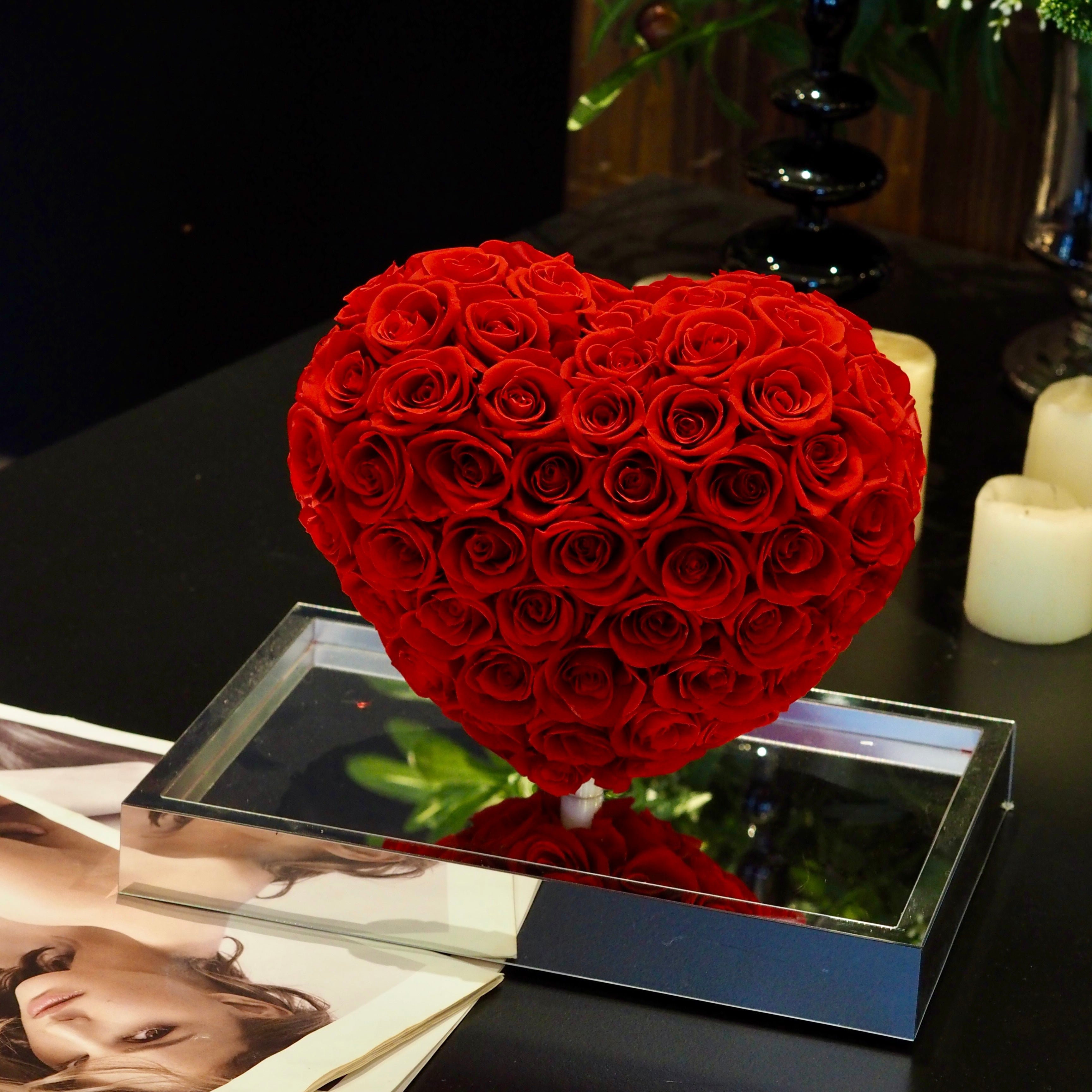 Heart-shaped arrangement of red roses on a reflective surface with a blurred background