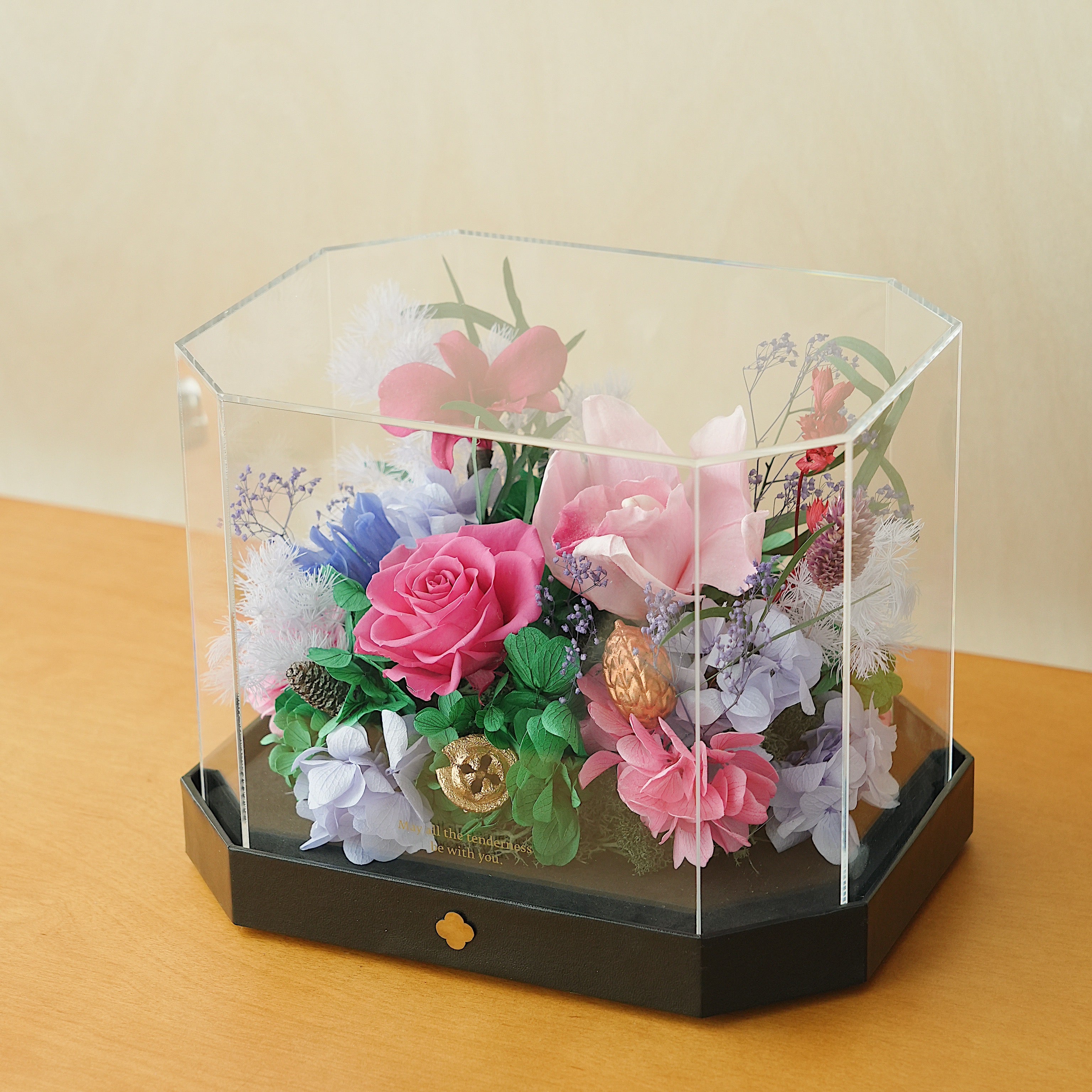 Decorative flower arrangement in a clear hexagonal glass display case on a wooden surface.