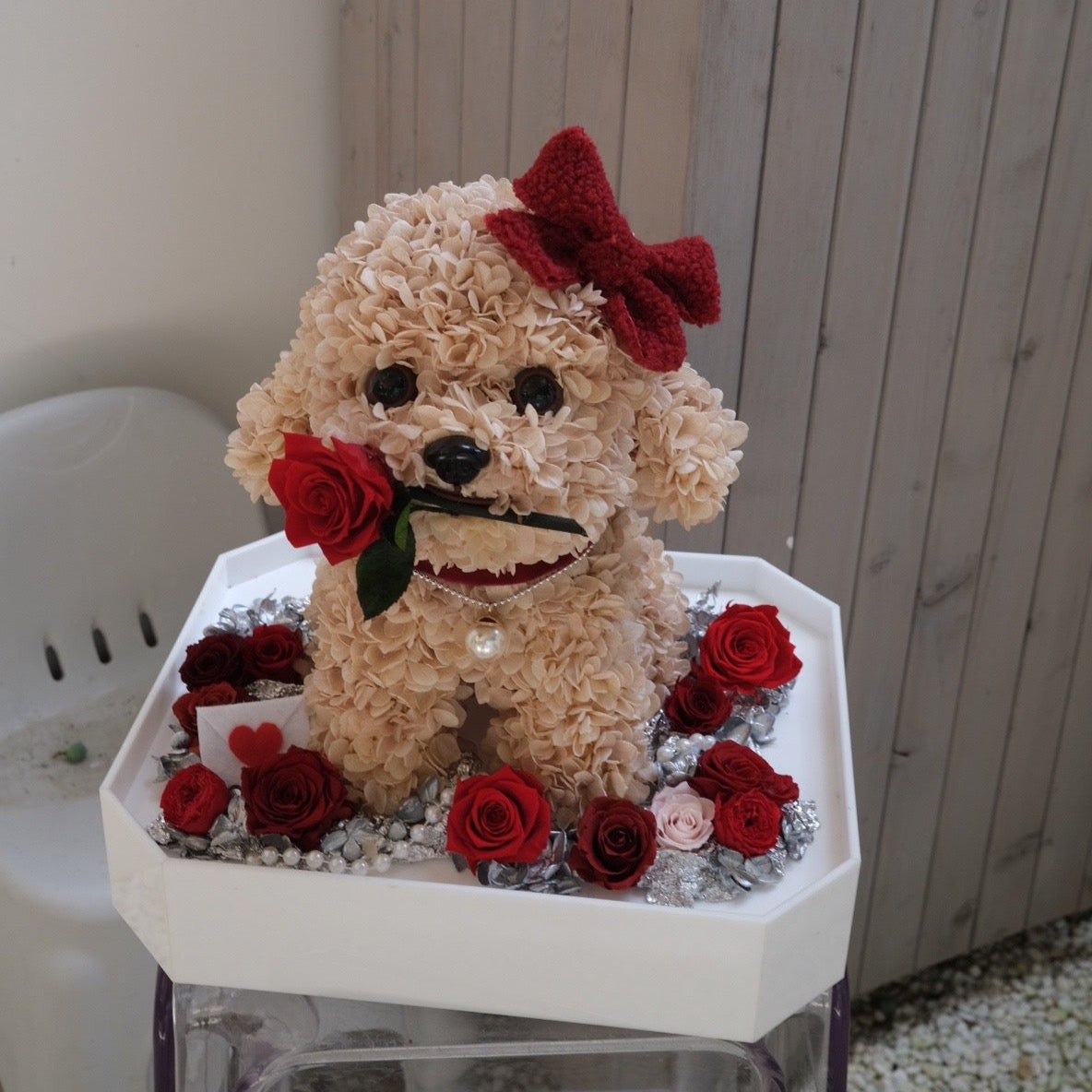 Floral Doggy Brown Poodle with red bow and roses on a small table against a wooden wall.