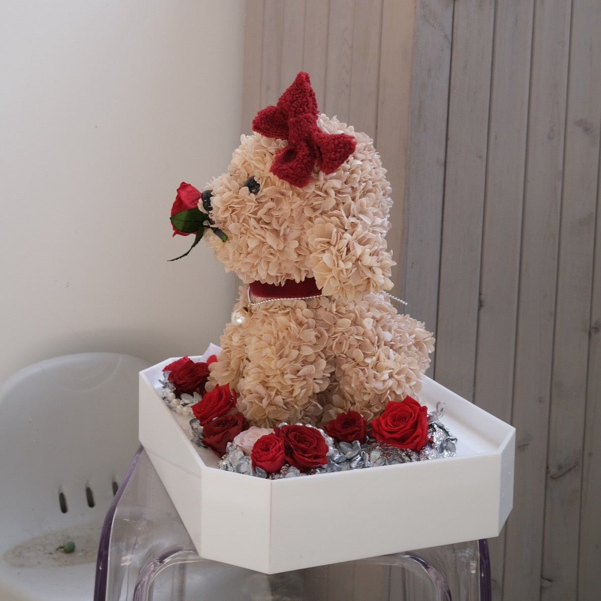 Floral Doggy Brown Poodle with red bow and flowers on a white stand against a wooden wall.