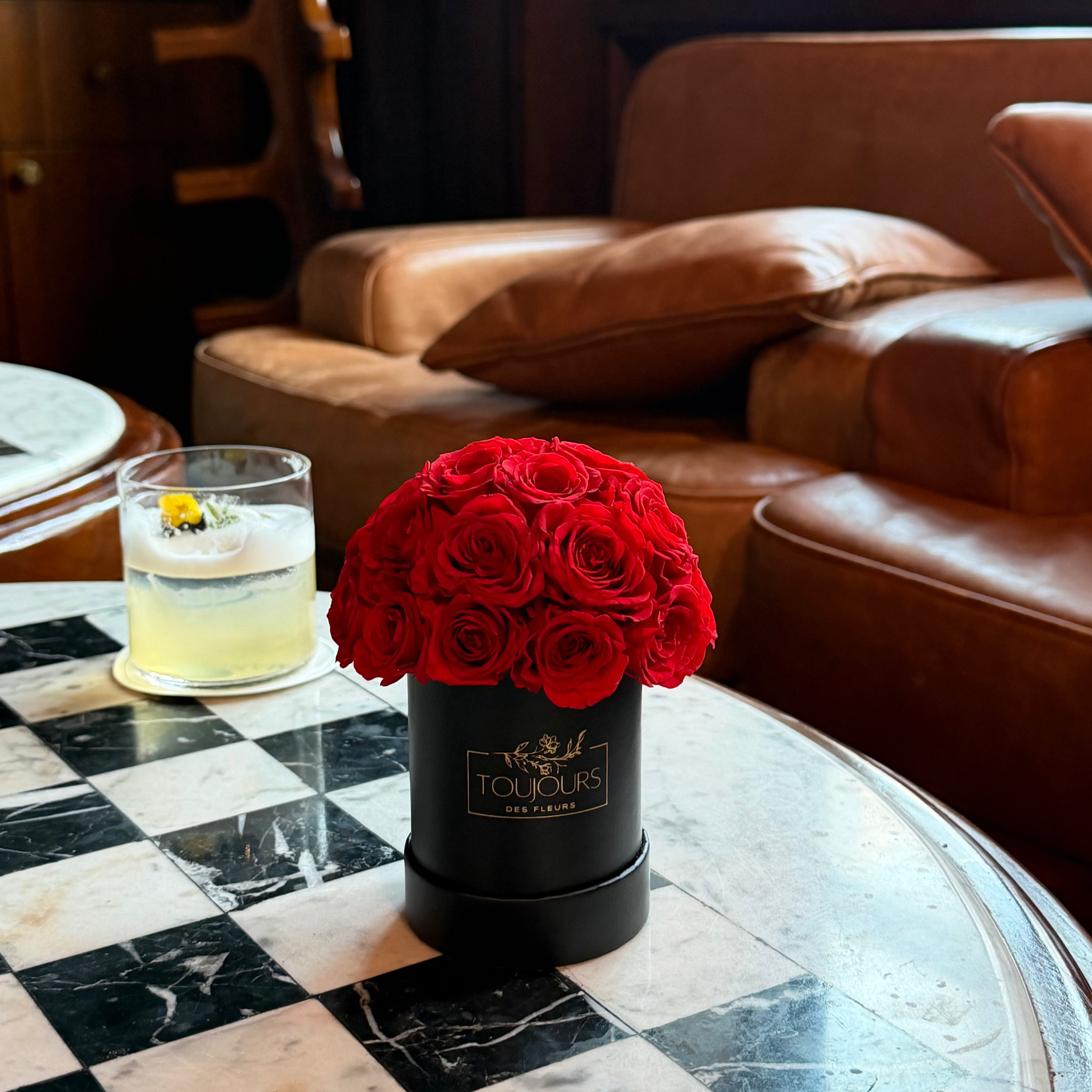 Red roses in a black container on a checkered table with a glass of a yellow drink, brown leather couch in the background.