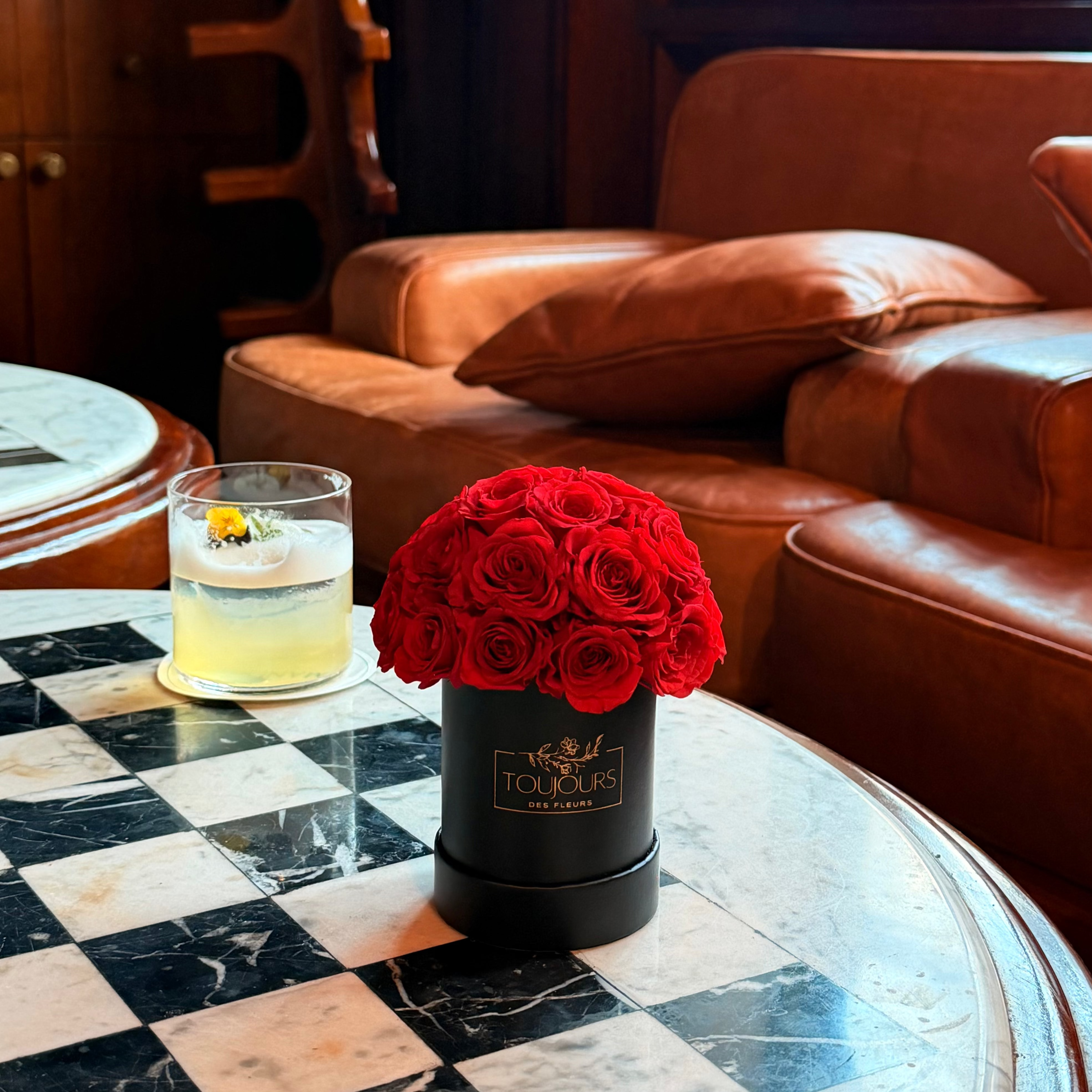 Red roses in a black container on a checkered table with a cocktail in a bar setting.