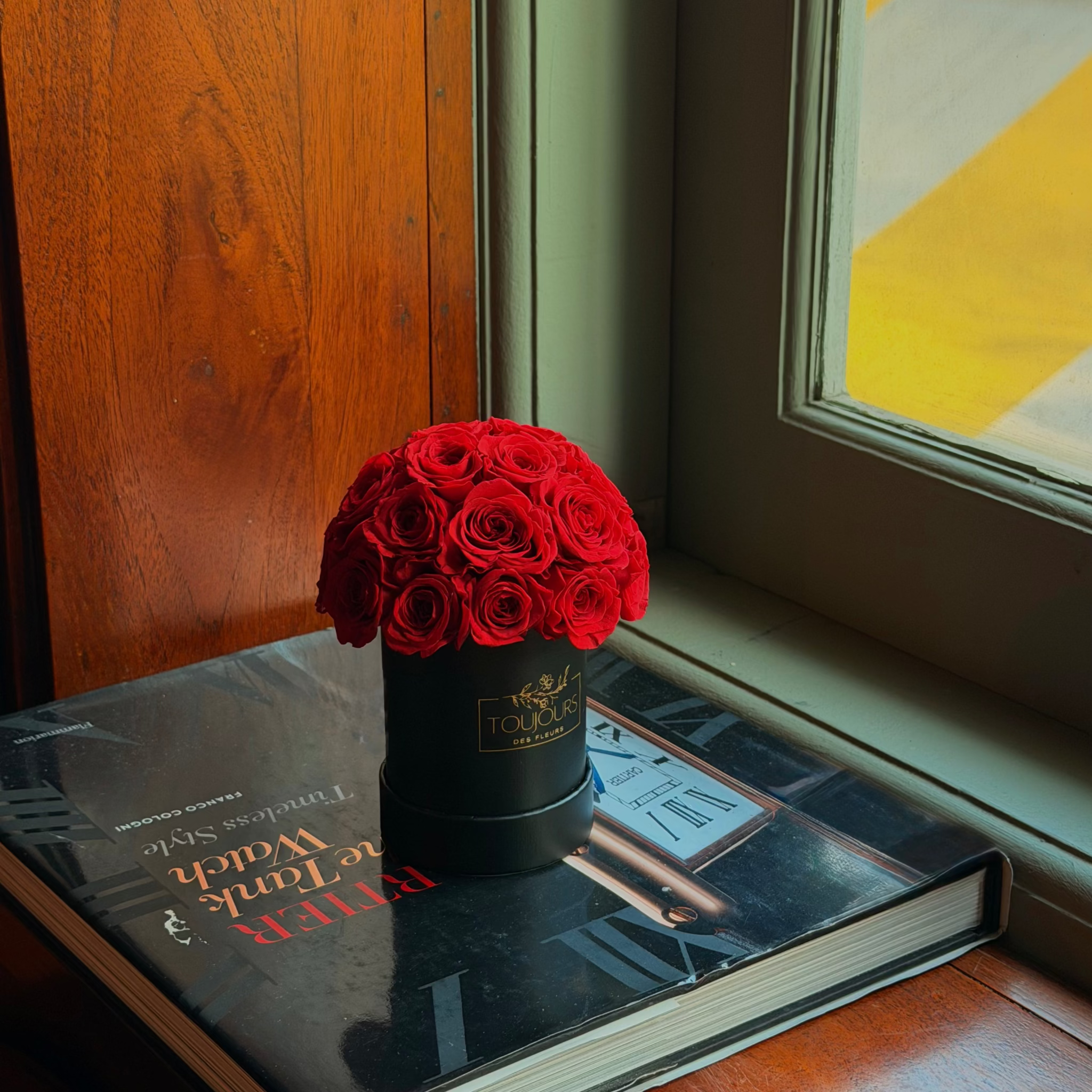 Red roses in a black container on a book next to a window