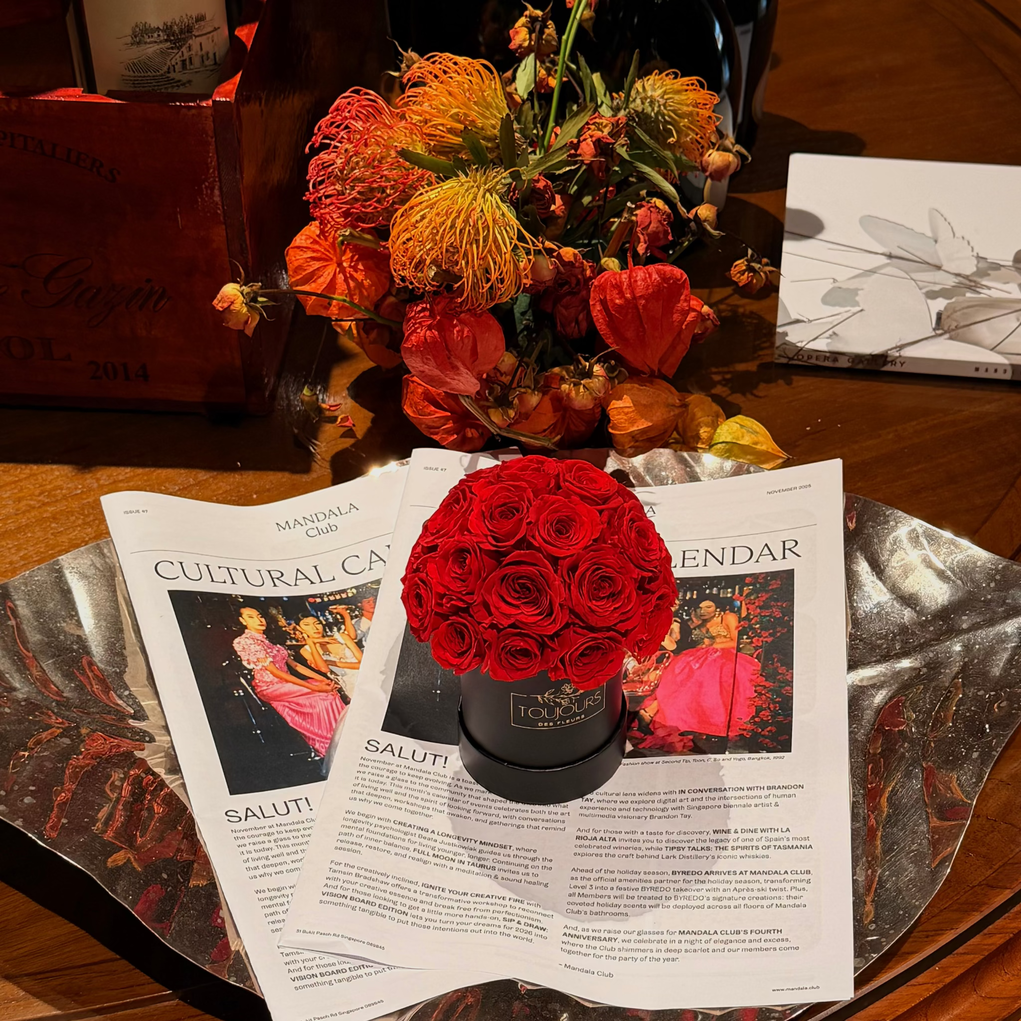 Decorative setup with red roses in a black box, flowers, and a magazine on a wooden table.