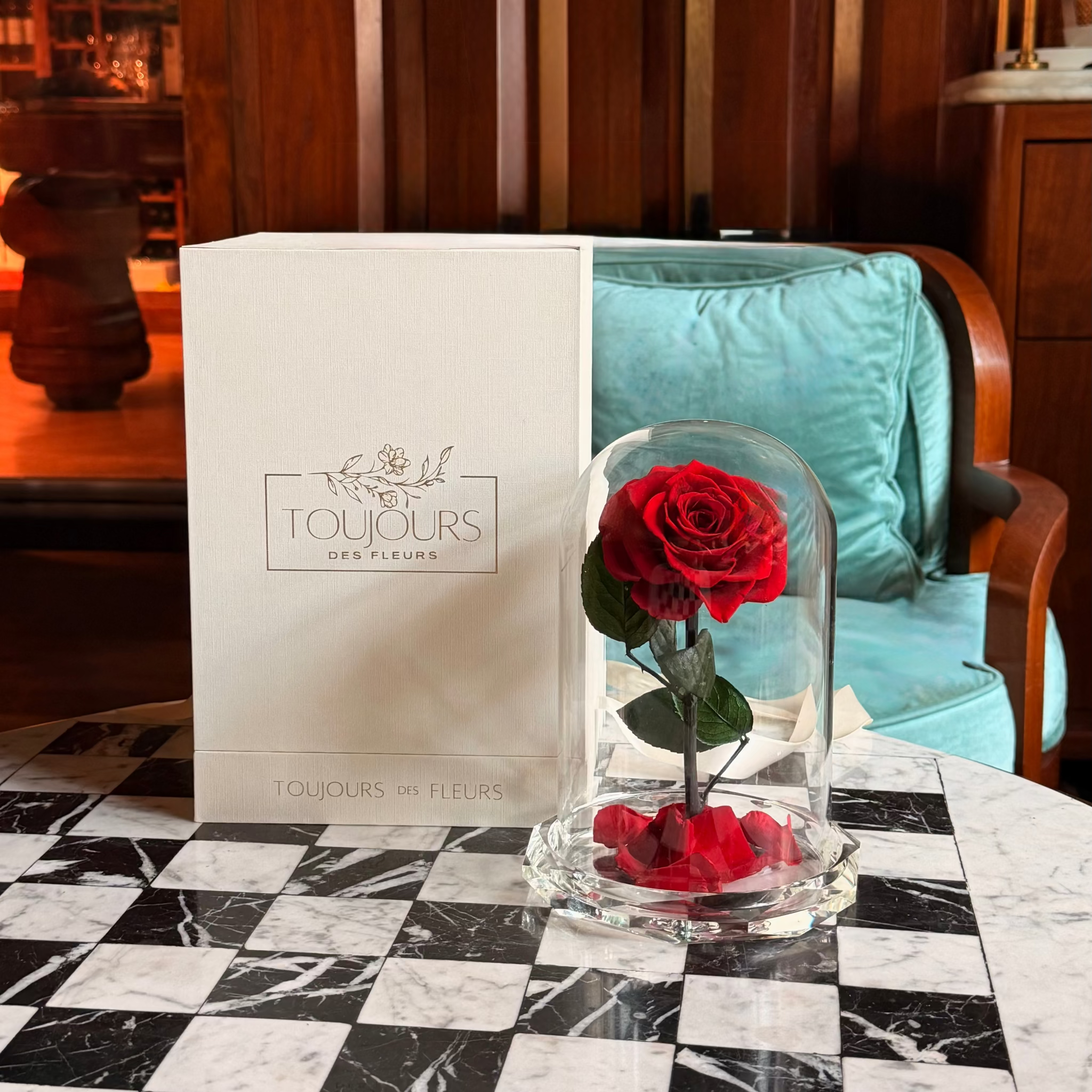 Red rose in a glass dome on a checkered floor with a box labeled ' Toujours des Fleurs' in the background.