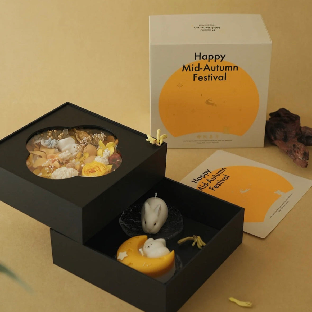 Flowers and candles in black box with 'Happy Mid-Autumn Festival' packaging on a beige background.