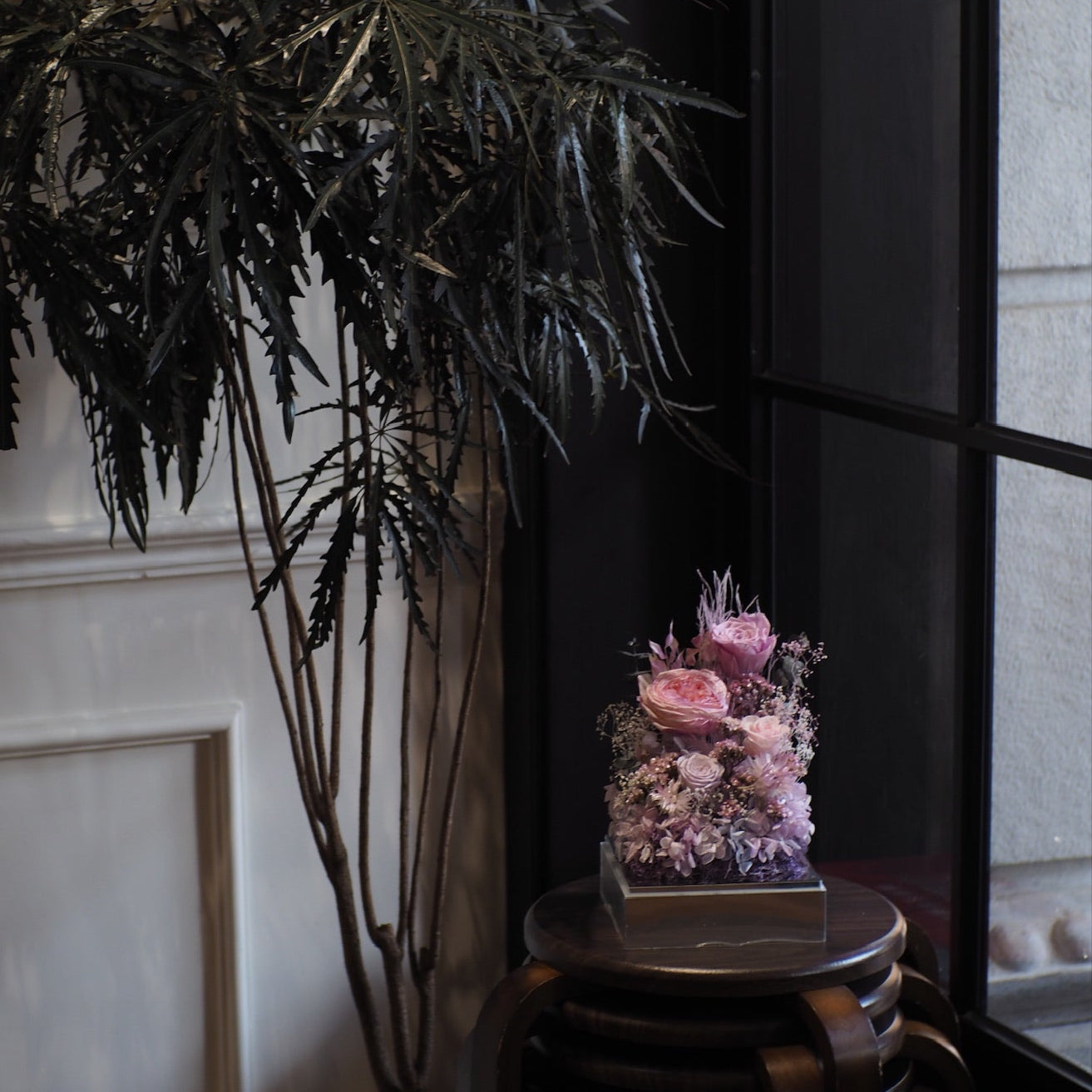 Potted plant next to a small floral arrangement on a wooden stool against a neutral wall.