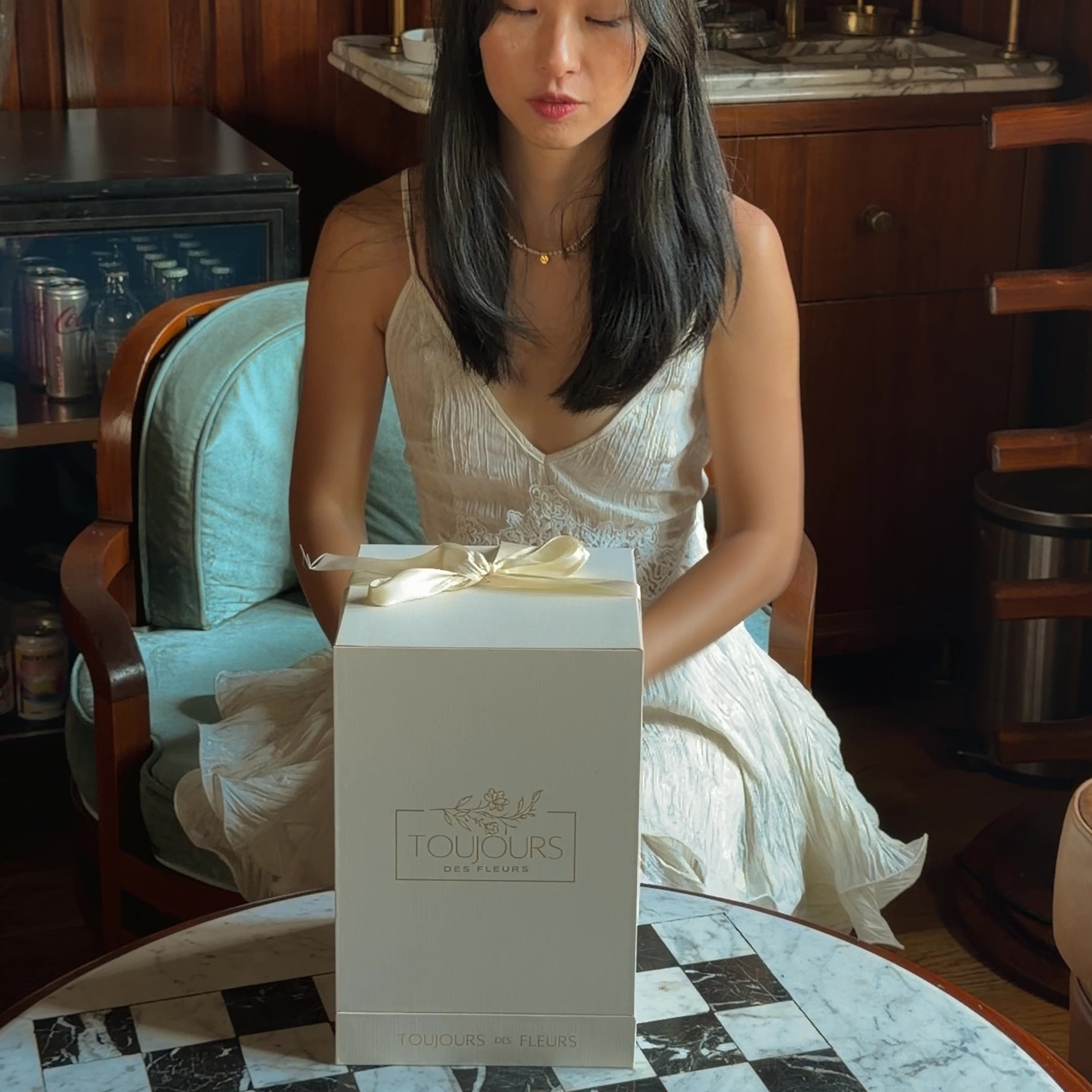 Red rose in a glass dome on a checkered floor with a box labeled ' Toujours des Fleurs' in the background.