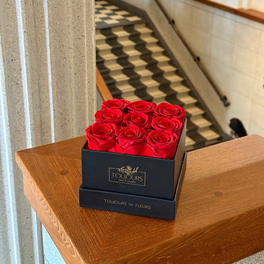 Black gift box of 9 preserved red roses from Toujours des Fleurs placed on a wooden ledge with vintage-style stairs behind